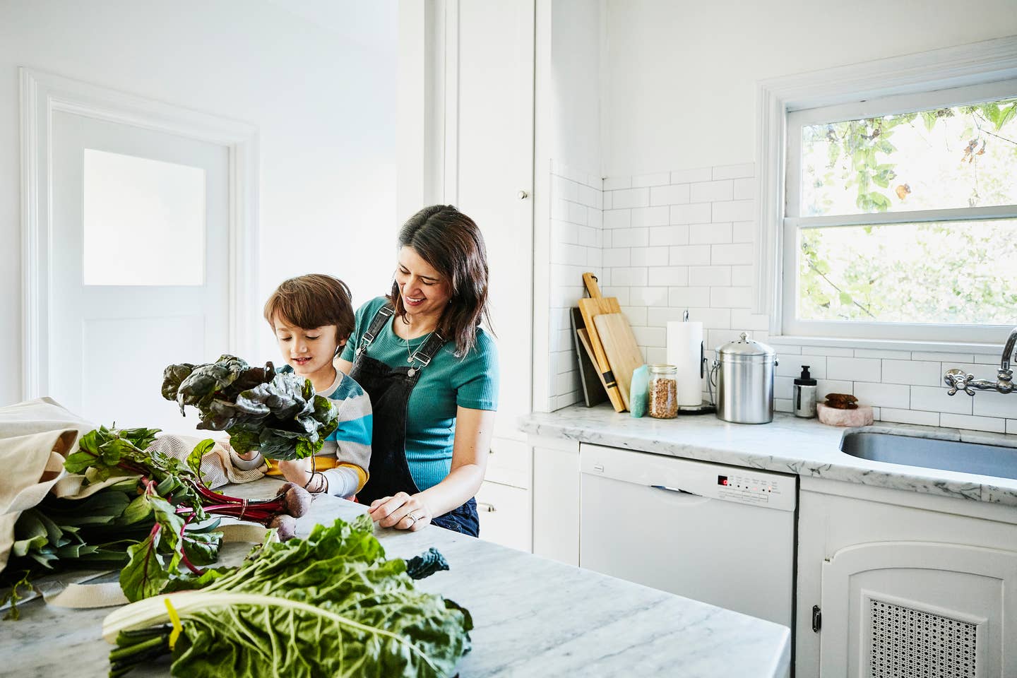 Smiling young son sitting in pregnant mothers lap while looking at fresh vegetables in kitchen after shopping